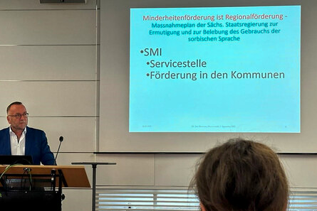 Mann hält Vortrag. Er steht am Rednerpult, hinter ihm hängt eine Leinwand, auf der eine Präsentation angezeigt wird.